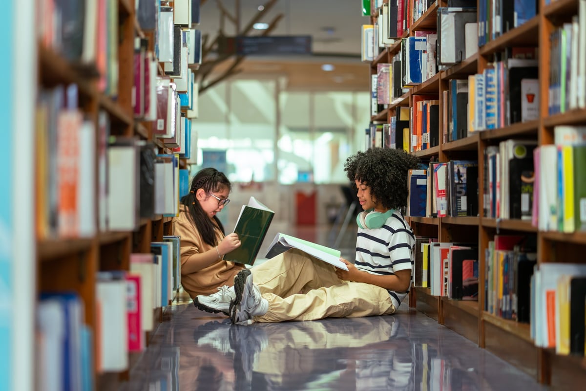 Student girls sitting together reading a book between the bookshelves in a school library